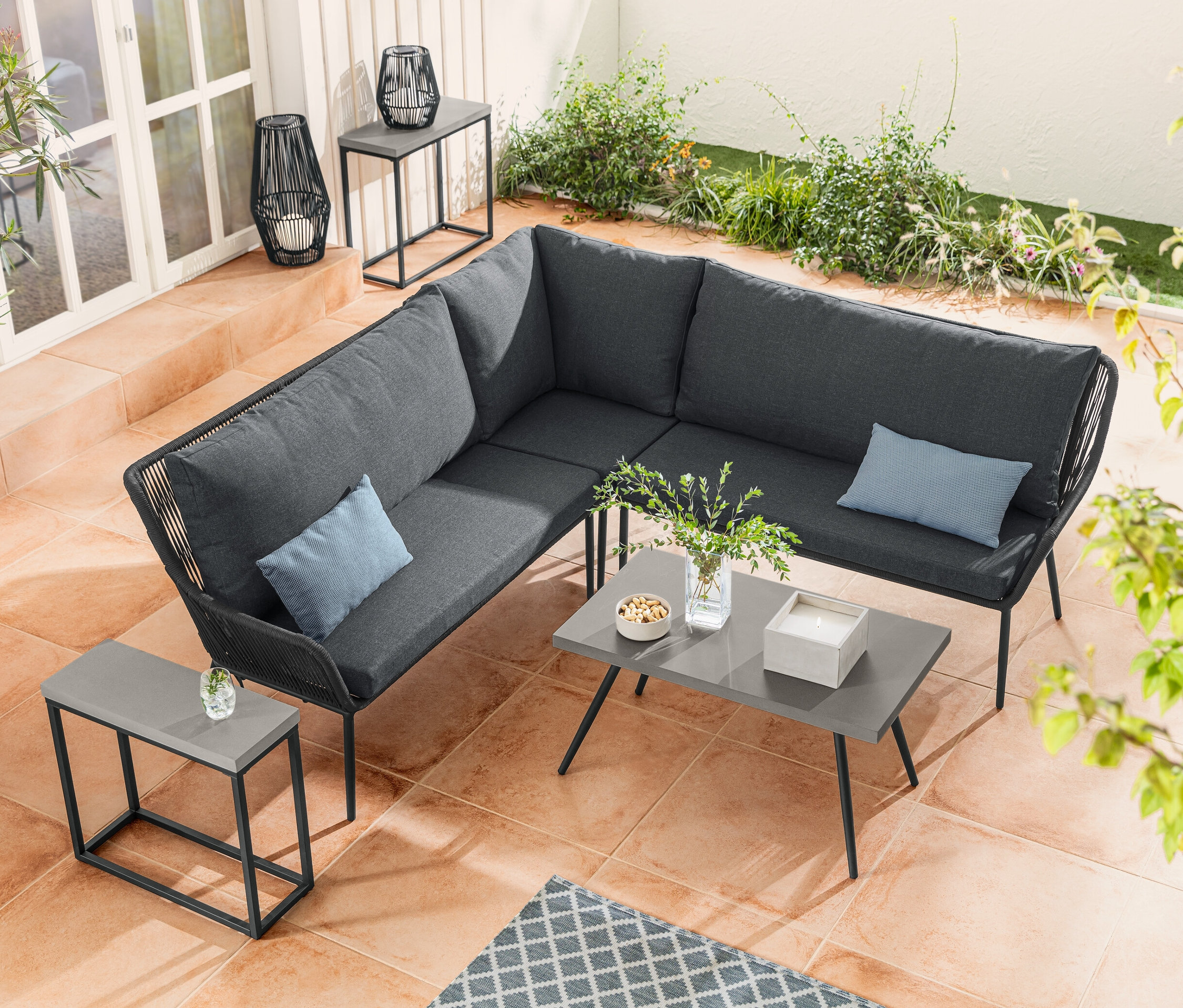 Un coin salon avec tressage textile est à côté d'une table basse avec plateau en polyciment. Un ensemble de tables console avec plateau en polyciment est placé à côté d'une lanterne solaire avec ruban en tissu.