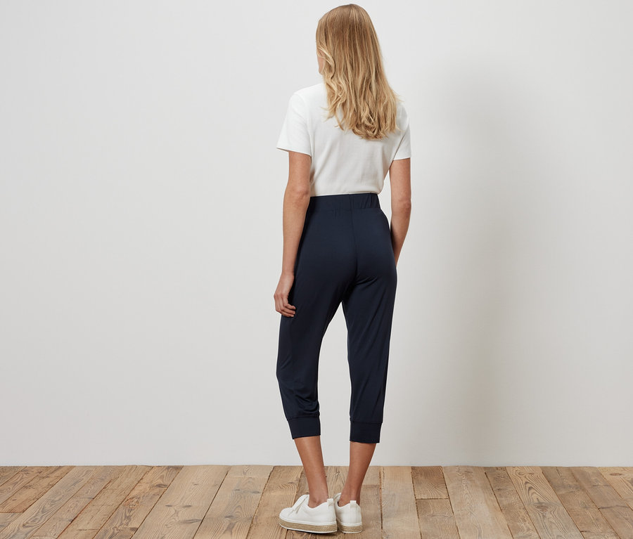 Une femme, vue de dos, porte un pantalon 3/4 en jersey bleu foncé, un t-shirt blanc et des baskets.