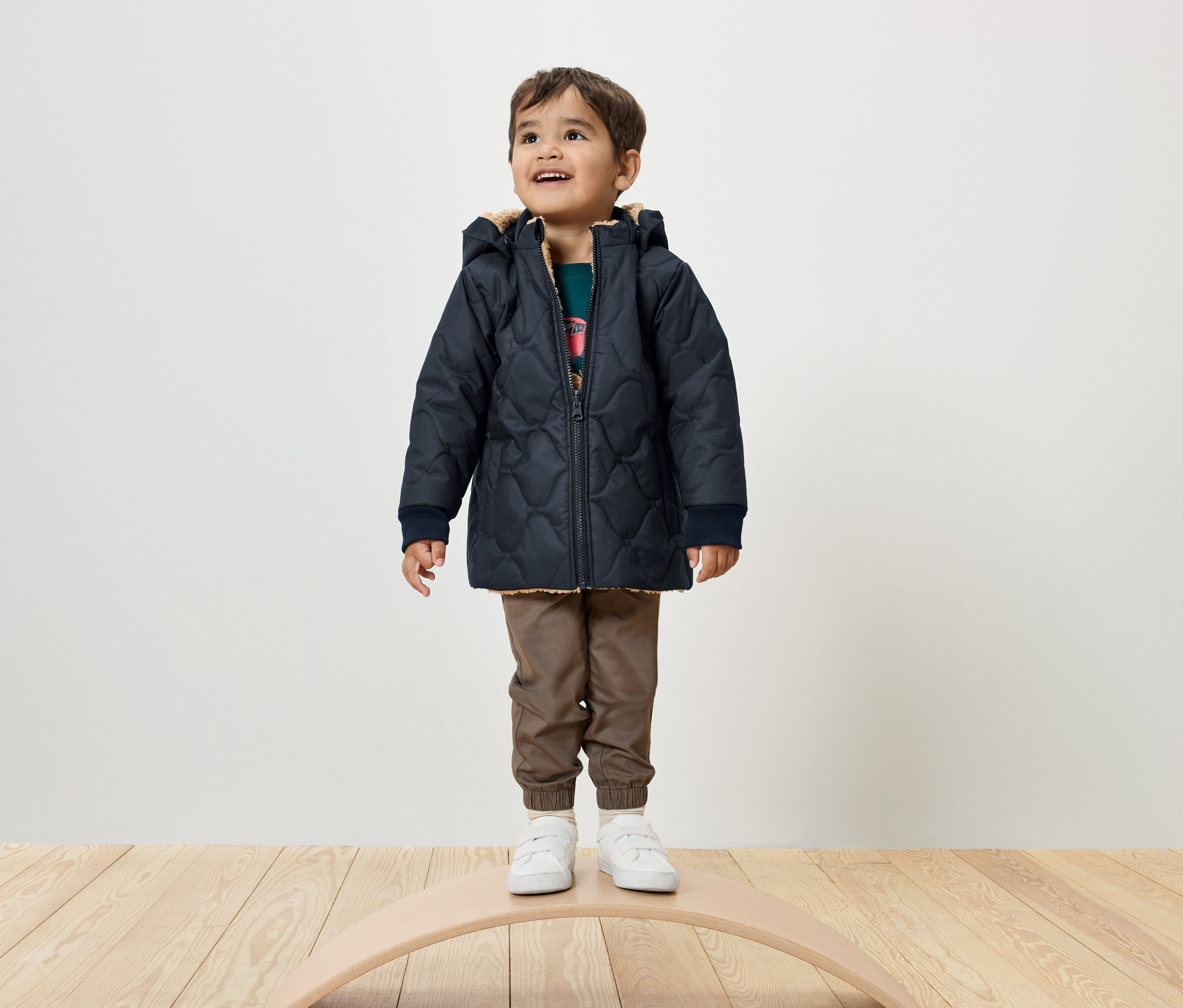 Un garçon se tient sur une planche d'équilibre, portant une veste matelassée bleu marine, un pantalon marron et des baskets blanches.