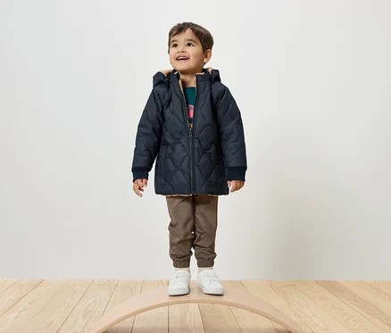 Un garçon se tient sur une planche d'équilibre, portant une veste matelassée bleu marine, un pantalon marron et des baskets blanches.
