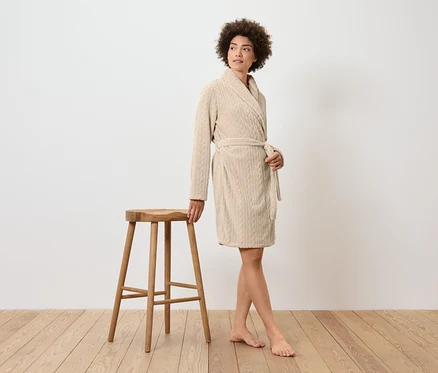 Femme en peignoir crème debout à côté d'un tabouret en bois.