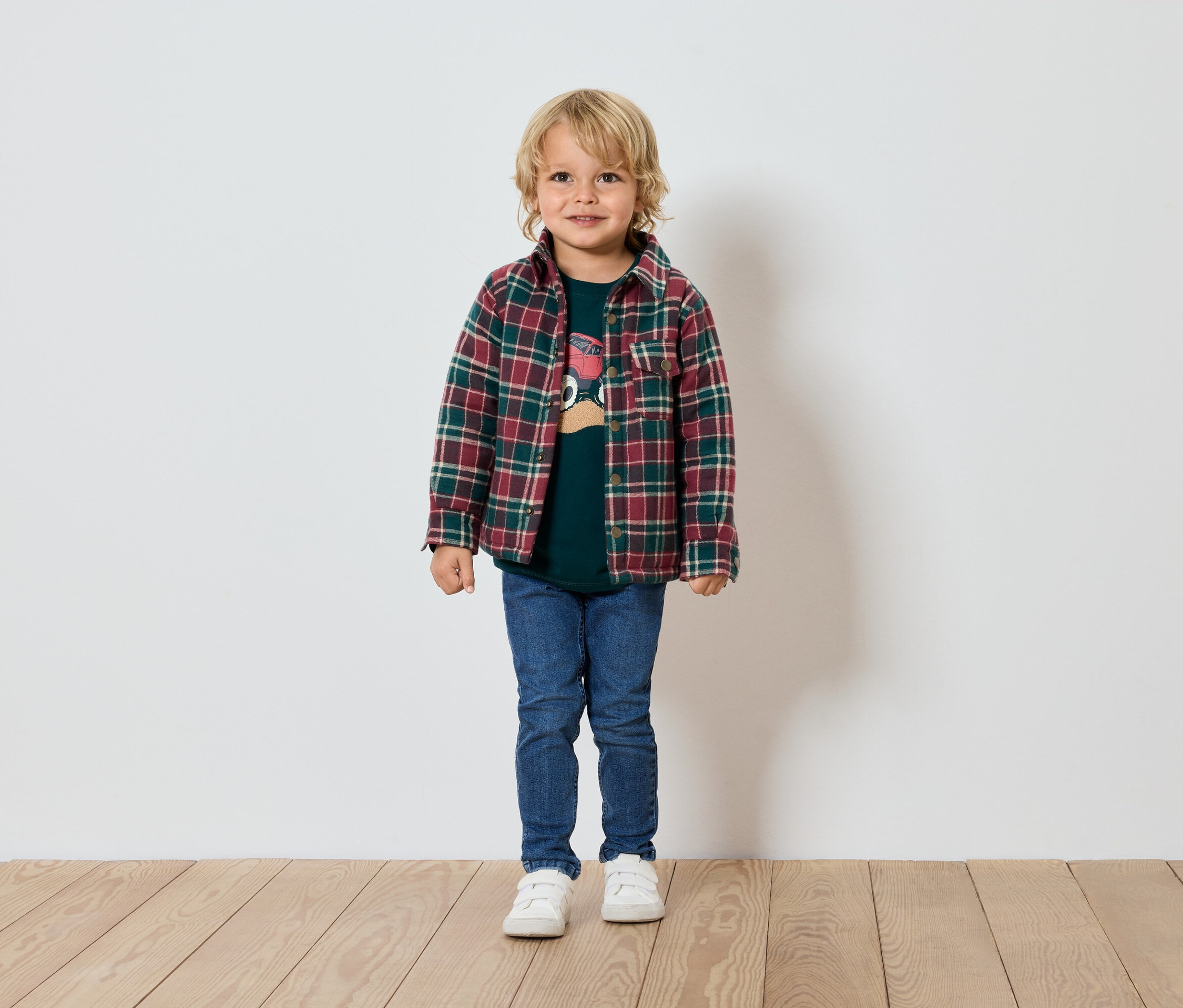Un garçon blond se tient devant un mur blanc, portant un t-shirt vert, une chemise à carreaux, un jean et des baskets.
