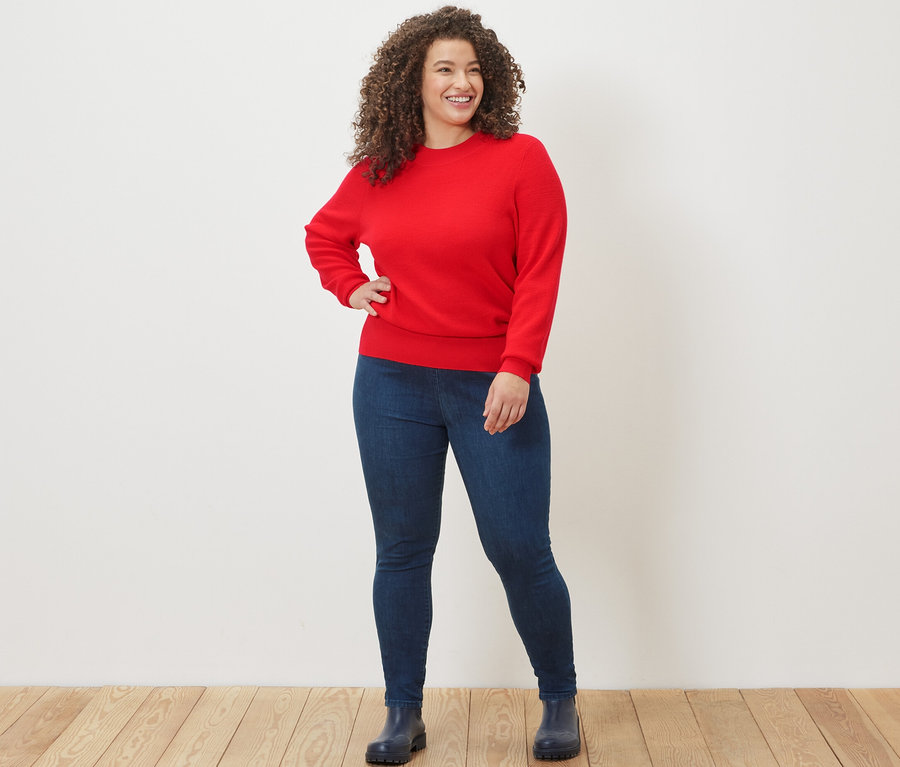 Une femme pose en pull en maille fine rouge et legging taille haute en denim.