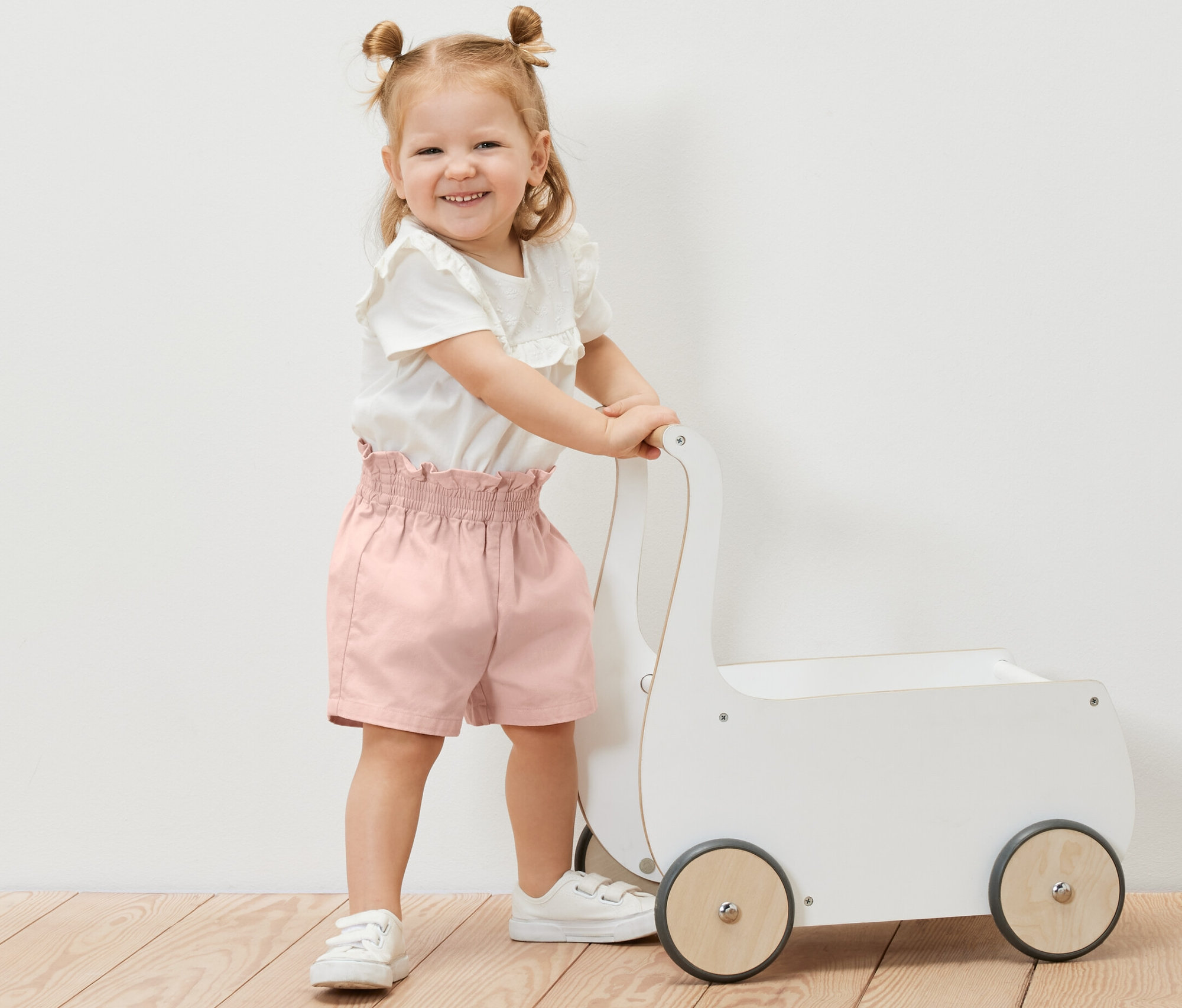 Une petite fille souriante pousse un chariot à jouets blanc et porte un short paper bag pour enfant.
