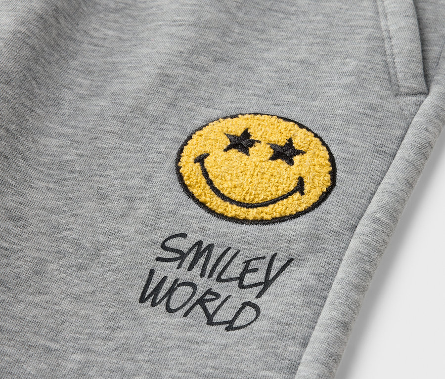 Détail d'un pantalon de jogging gris pour enfant SMILEYWORLD® avec un patch smiley jaune.