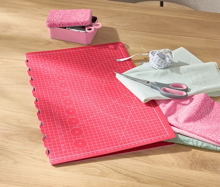 Un tapis de découpe et de bricolage pliable rose est posé sur une table à côté d'une boîte rose contenant des ciseaux, des tissus et un mètre ruban.