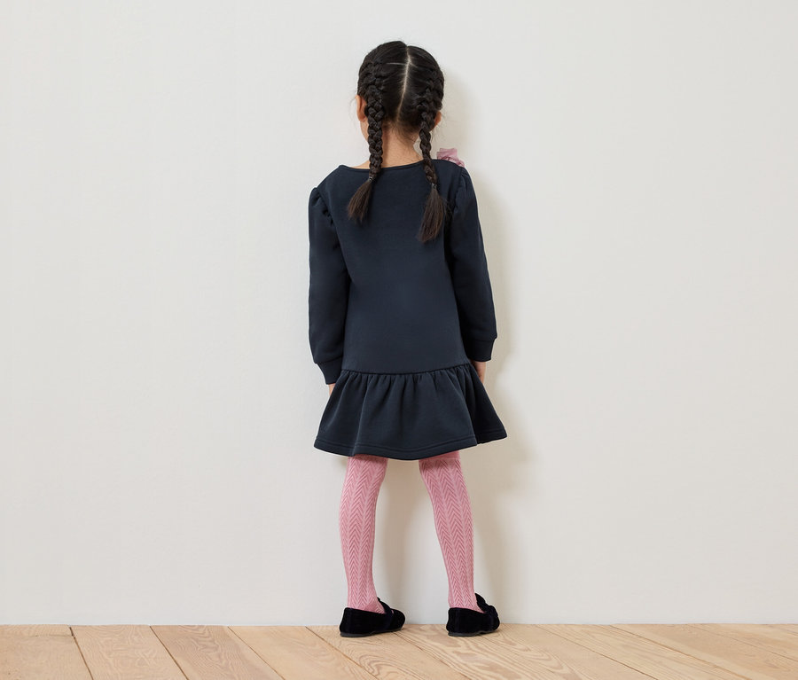 Une fillette avec des tresses se tient dos à la caméra, portant une robe bleue, des collants roses à motifs et des chaussures noires.