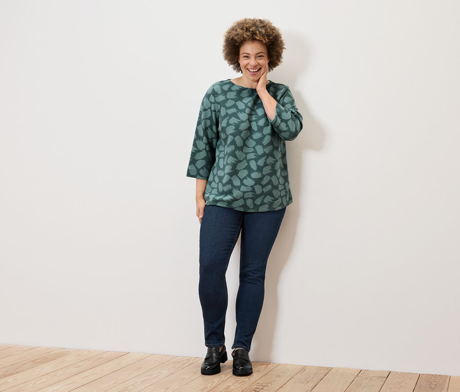 Une femme souriante aux cheveux bouclés pose dans un haut vert imprimé, un jean et des chaussures noires.