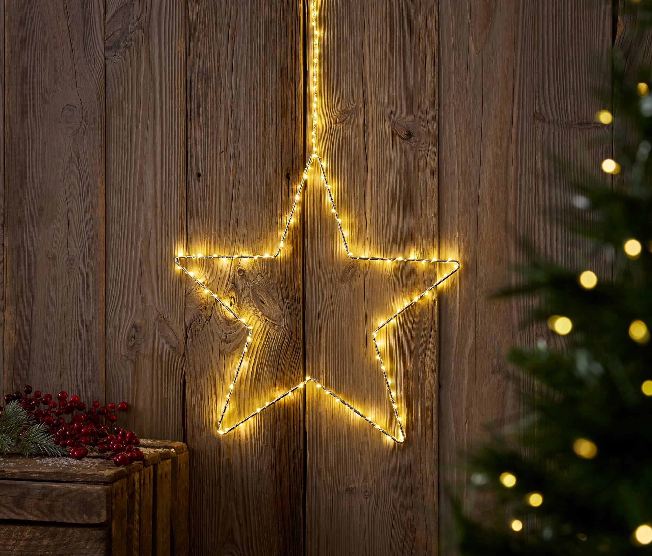 Une étoile lumineuse jaune est accrochée à un mur en bois à côté d'un arbre de Noël et d'une boîte en bois avec des baies rouges.