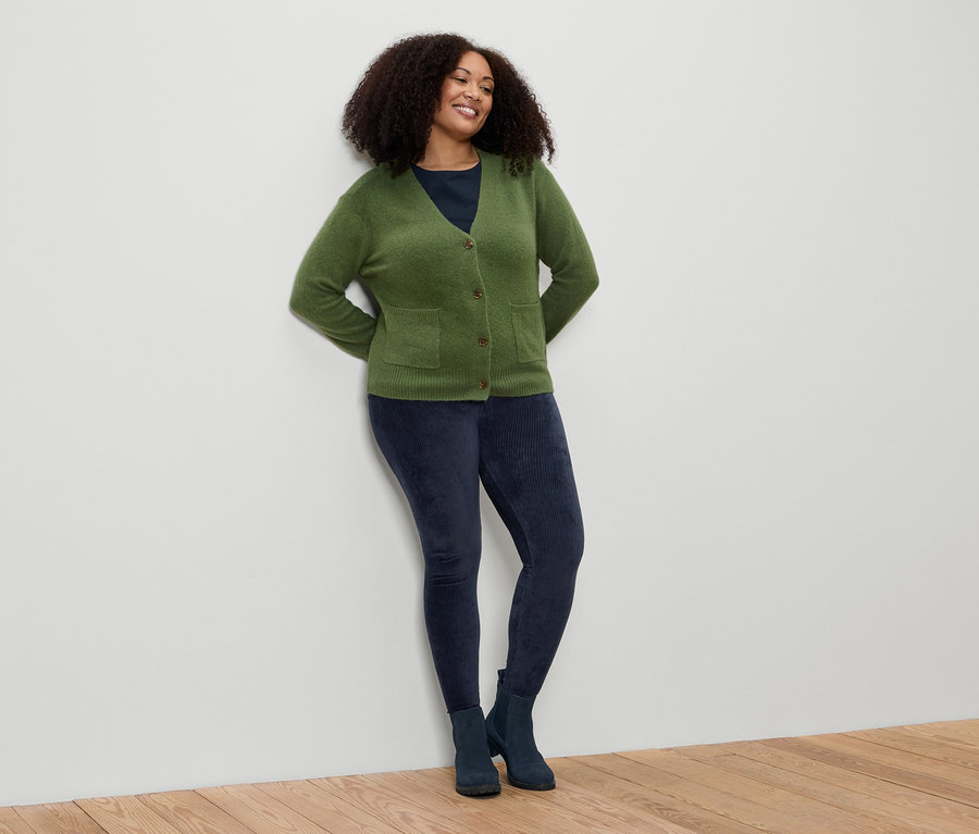 Une femme aux cheveux bouclés s'appuie contre un mur. Elle porte un cardigan vert, un legging bleu et des bottes.