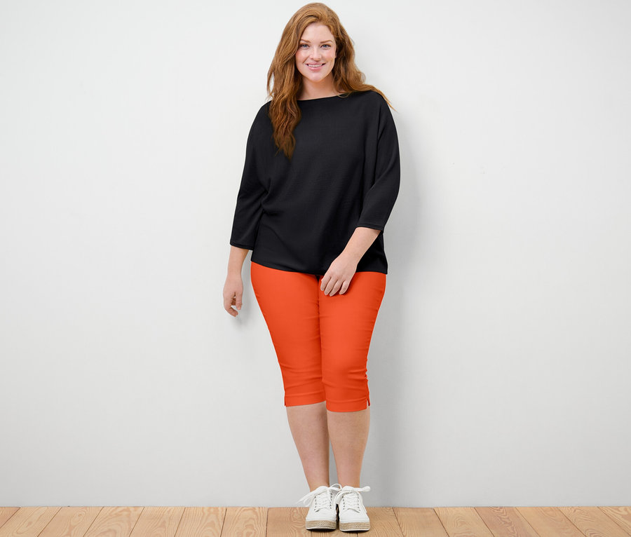 Une femme se tient debout et porte un pantalon 3/4 stretch orange et un pull en maille fine noir.