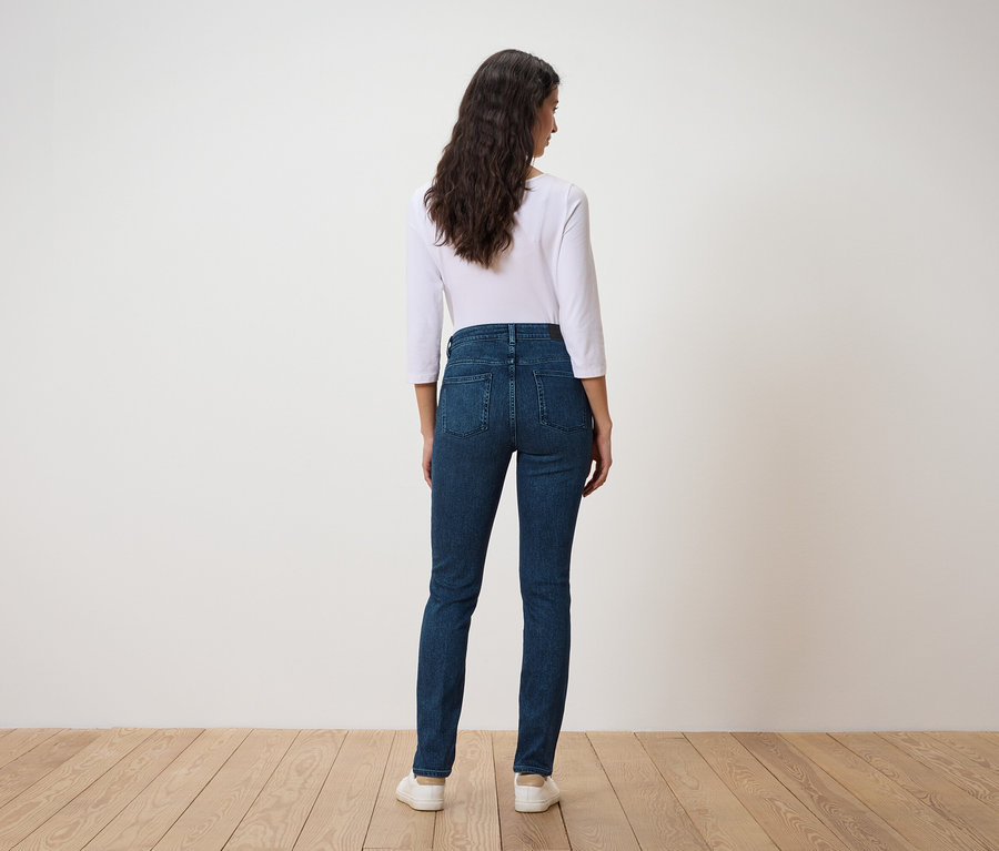 Une femme aux cheveux ondulés foncés se tient dos à l'appareil photo, portant un t-shirt blanc à manches trois-quarts et un jean bleu foncé.