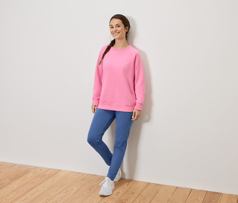 Femme aux cheveux tressés portant un sweat-shirt rose, un pantalon bleu et des chaussures blanches, appuyée contre un mur.