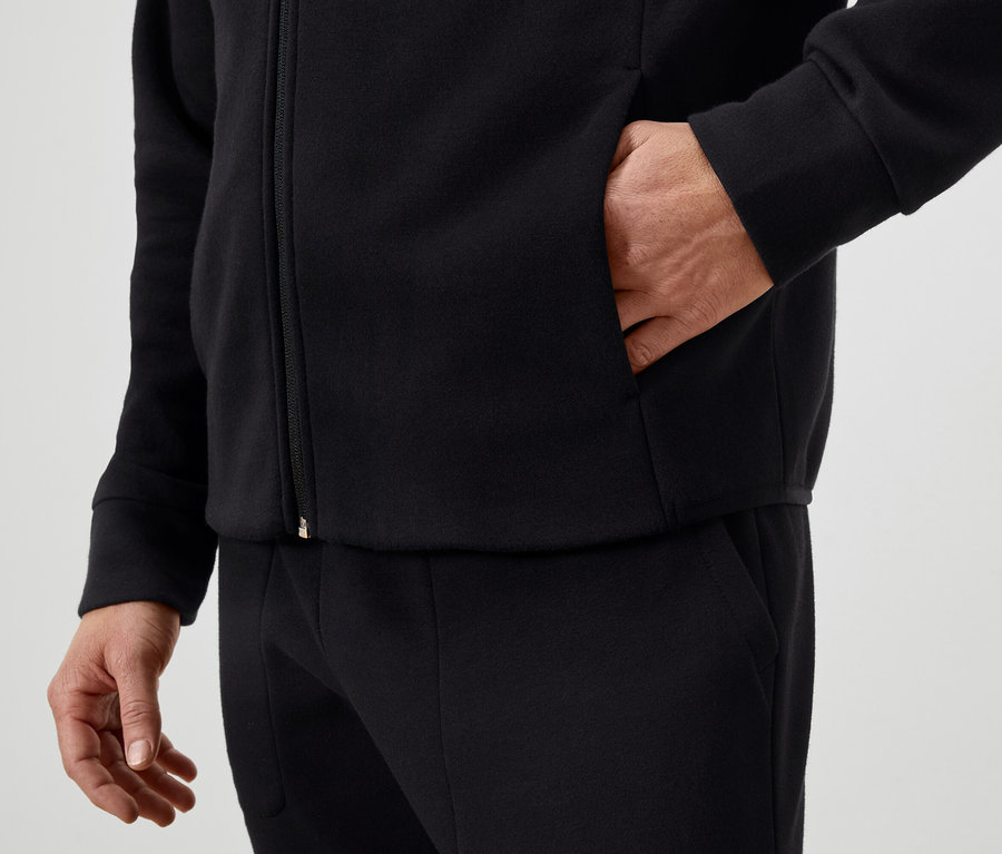 Un homme a la main dans la poche d'un blouson noir zippé et porte un pantalon noir.