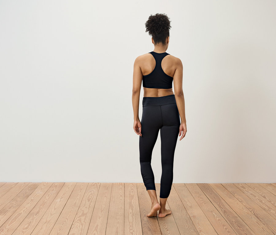 Une femme en tenue de sport noire se tient dos à la caméra sur un plancher en bois.