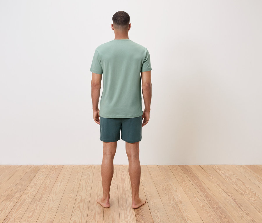 Un homme se tient pieds nus, dos au spectateur, portant un t-shirt vert et un short.