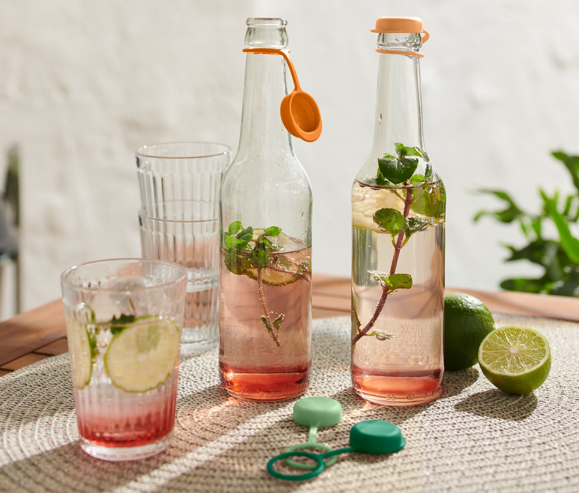 Deux bouteilles en verre avec boisson, deux verres, des limes et 4 bouchons de bouteille sur une table.