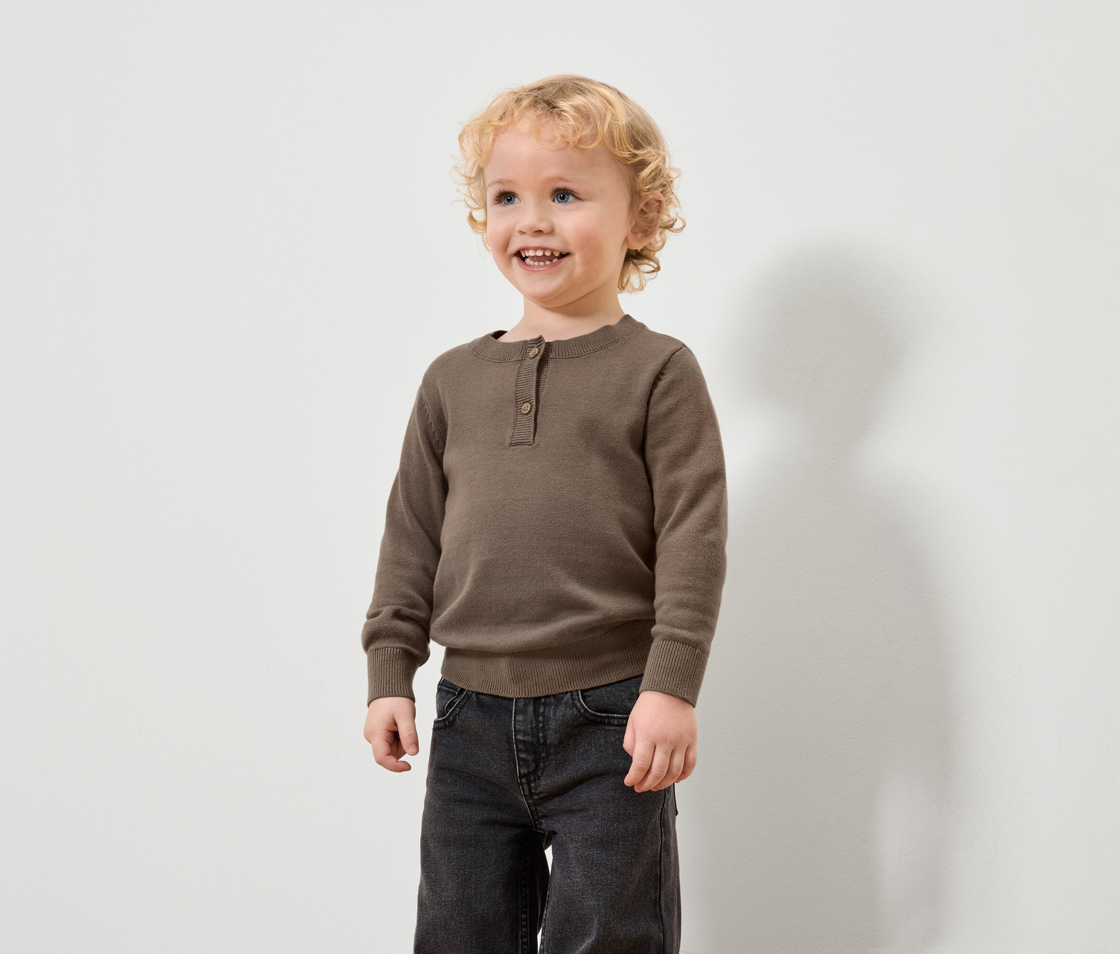 Un garçon aux cheveux blonds bouclés porte un pull marron et un jean noir.