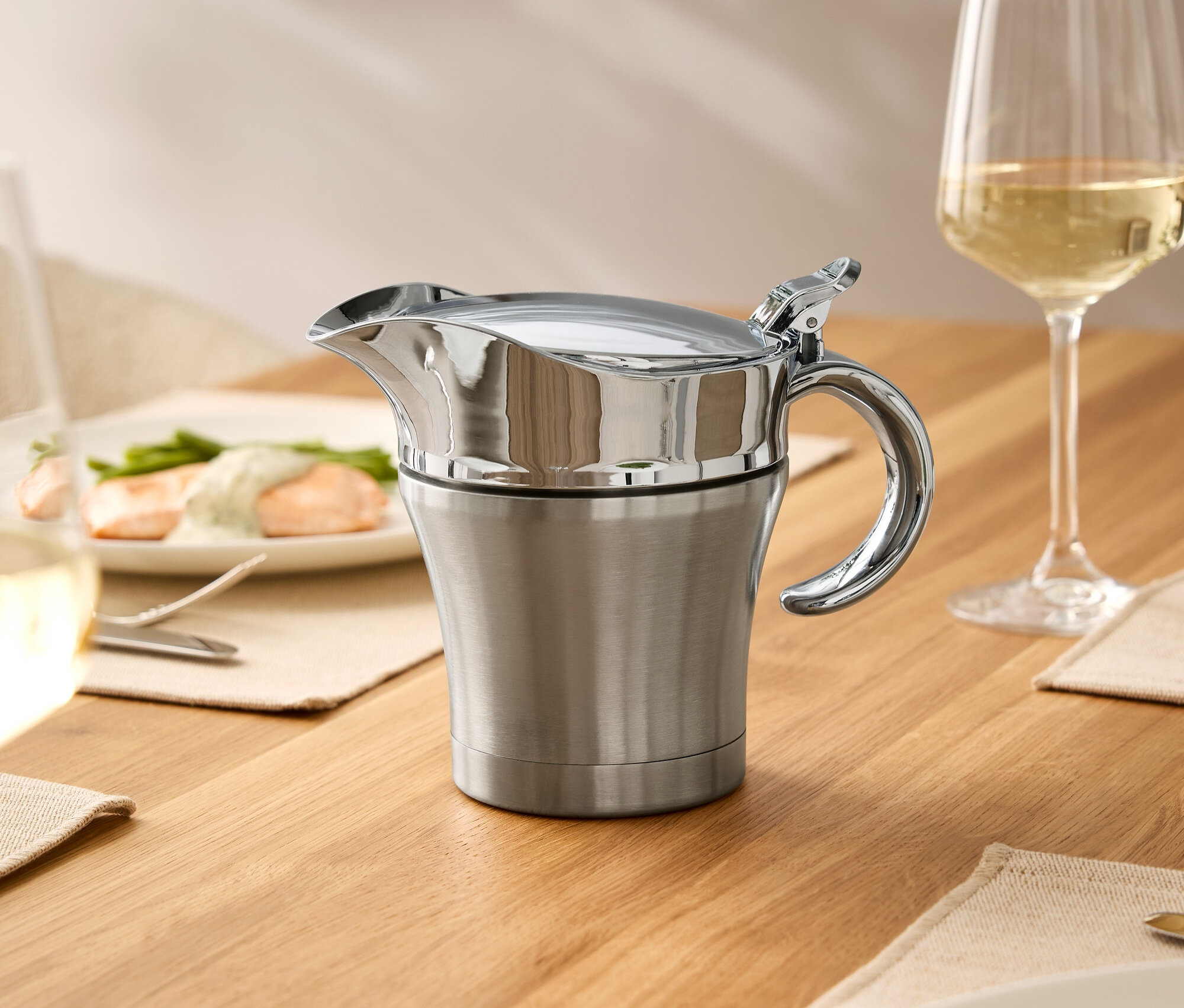 Sur une table en bois se trouvent une saucière en acier inoxydable avec couvercle, un verre de vin et des assiettes de nourriture.