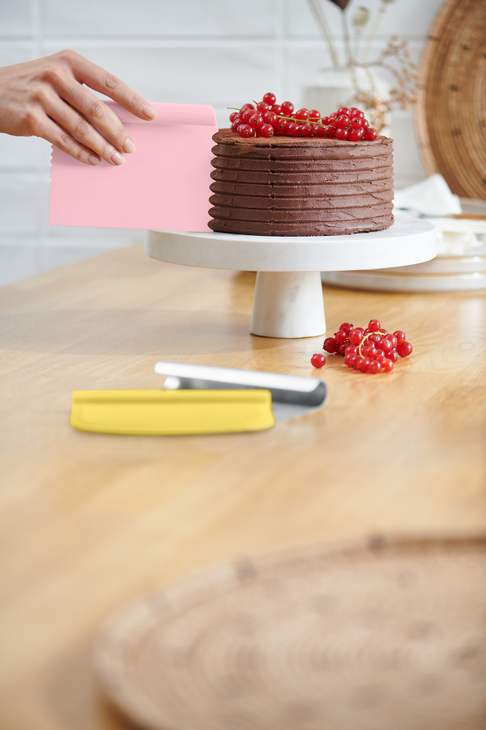 Une main lisse le glaçage d'un gâteau au chocolat avec une spatule rose. Le gâteau repose sur un présentoir blanc et est décoré de groseilles rouges. Une spatule jaune est posée sur la table.