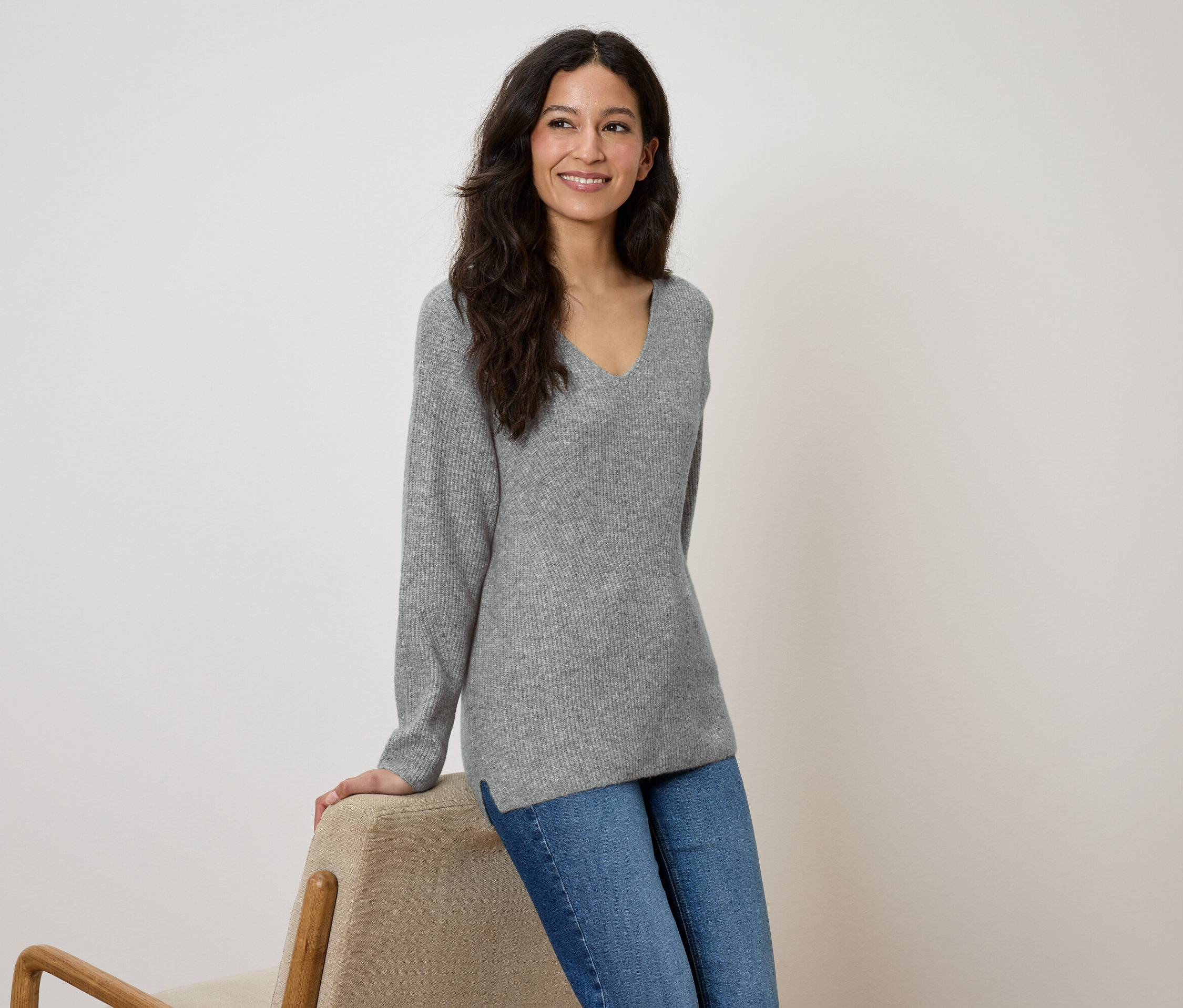 Une femme souriante, vêtue d'un pull gris et d'un jean bleu, s'appuie sur une chaise beige.