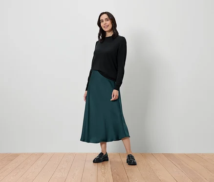 Une femme se tient debout et pose en pull noir et jupe vert foncé.