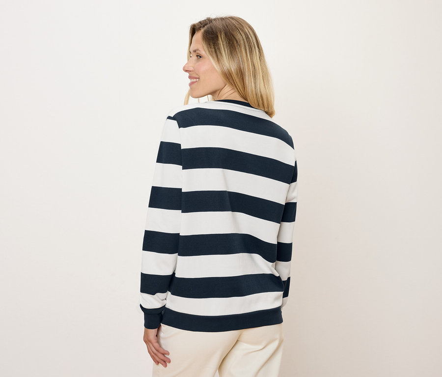 Une femme regarde par-dessus son épaule et porte un t-shirt à manches longues rayé bleu et blanc et un pantalon.