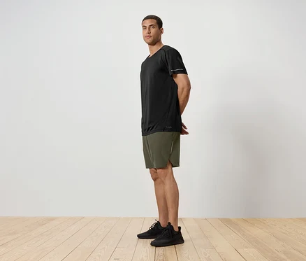 Vue de côté d'un homme portant un t-shirt noir, un short vert et des baskets noires debout sur un plancher en bois devant un mur blanc.