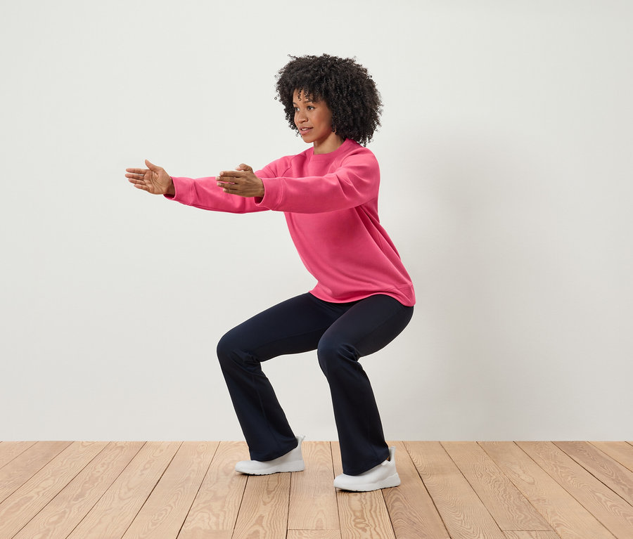 Femme en sweat-shirt rose et pantalon bleu marine faisant des squats, les bras tendus.