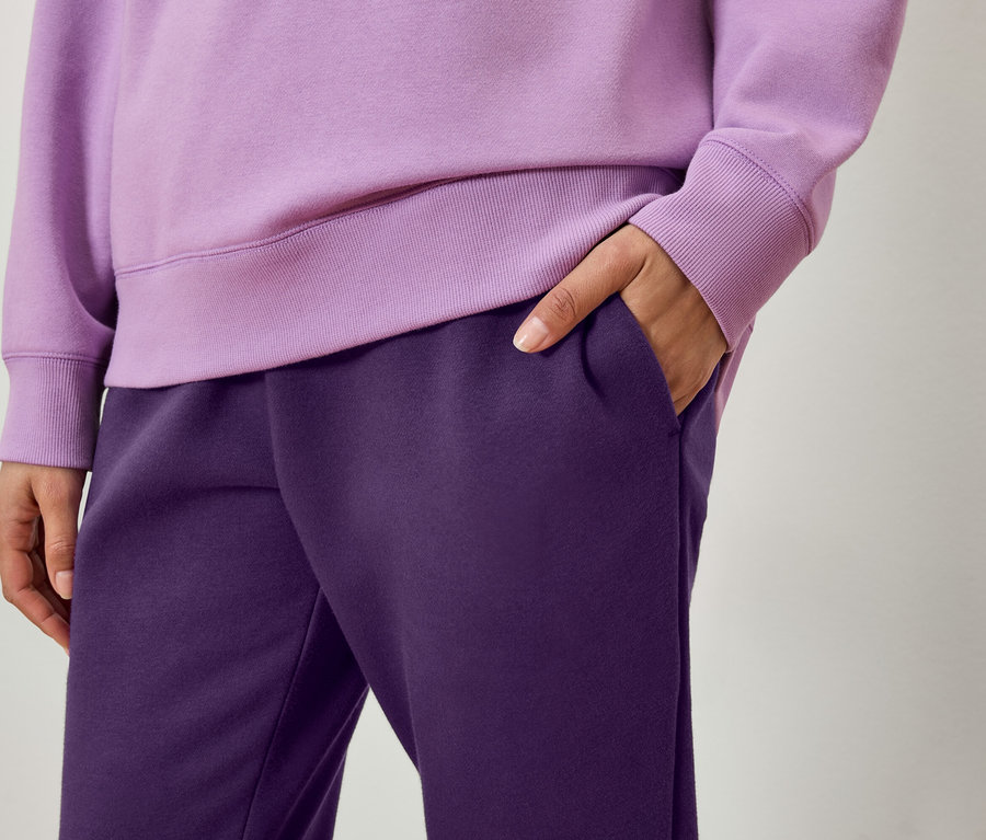 Gros plan d'une personne portant un sweat-shirt violet et un pantalon de survêtement violet foncé, une main dans la poche.