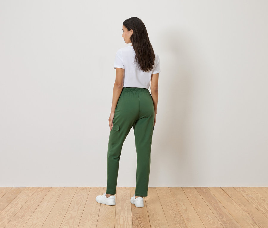 Une femme se tient dos à la caméra, portant un t-shirt blanc, un pantalon vert et des baskets blanches.