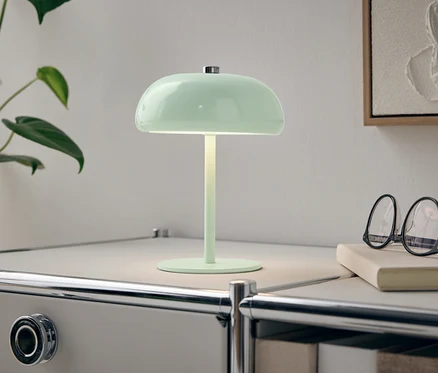 Une lampe verte est posée sur une table de chevet blanche à côté d'une plante et de lunettes.