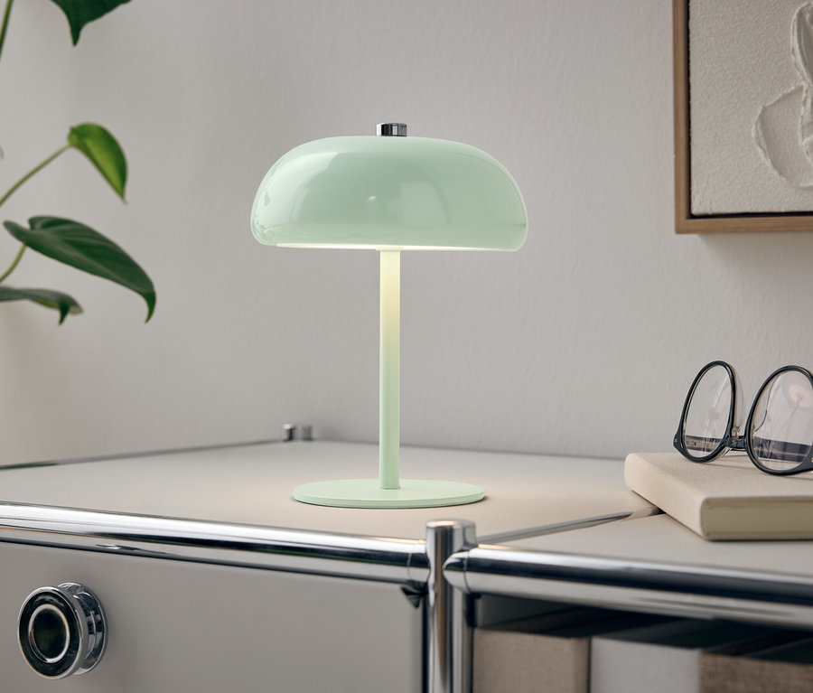 Une lampe verte est posée sur une table de chevet blanche à côté d'une plante et de lunettes.