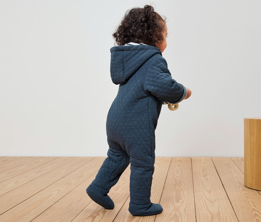Un bébé en combinaison matelassée bleue avec capuche marche sur un plancher en bois.