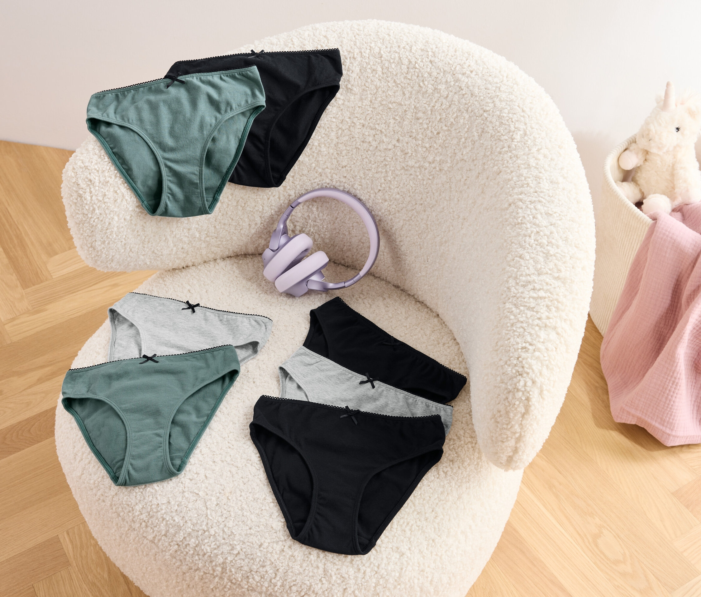 Six slips de couleur verte, grise et noire et un casque violet sont posés sur un fauteuil en tissu bouclé. Dans un panier, on trouve une couverture rose et une peluche de licorne.
