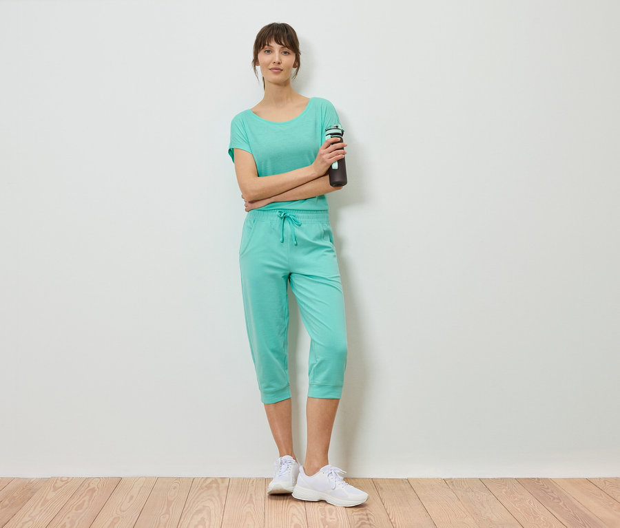 Femme portant un t-shirt menthe et un pantacourt, debout, les bras croisés, tenant une bouteille d'eau.