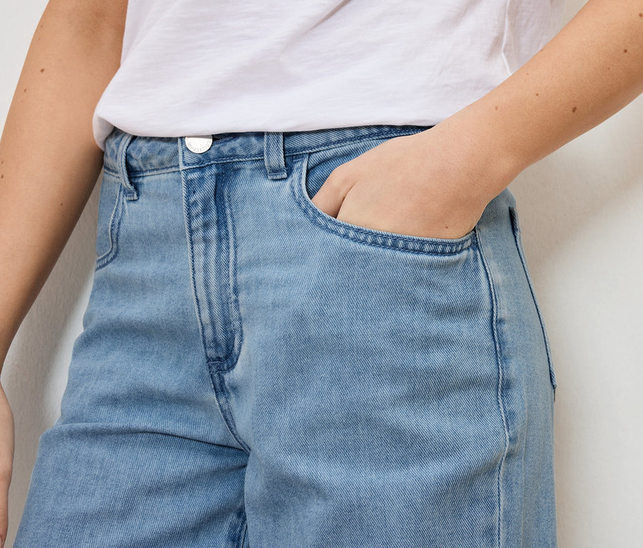 Gros plan d'une femme portant un jean bleu clair et un t-shirt blanc, avec sa main dans sa poche.