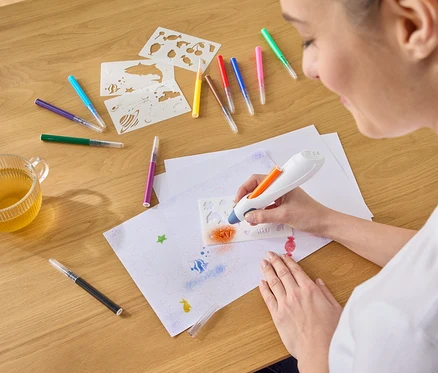Une femme pulvérise de la peinture orange à l'aide d'un aérographe à travers un pochoir sur une feuille de papier, entourée de stylos, de pochoirs et d'une tasse de thé.