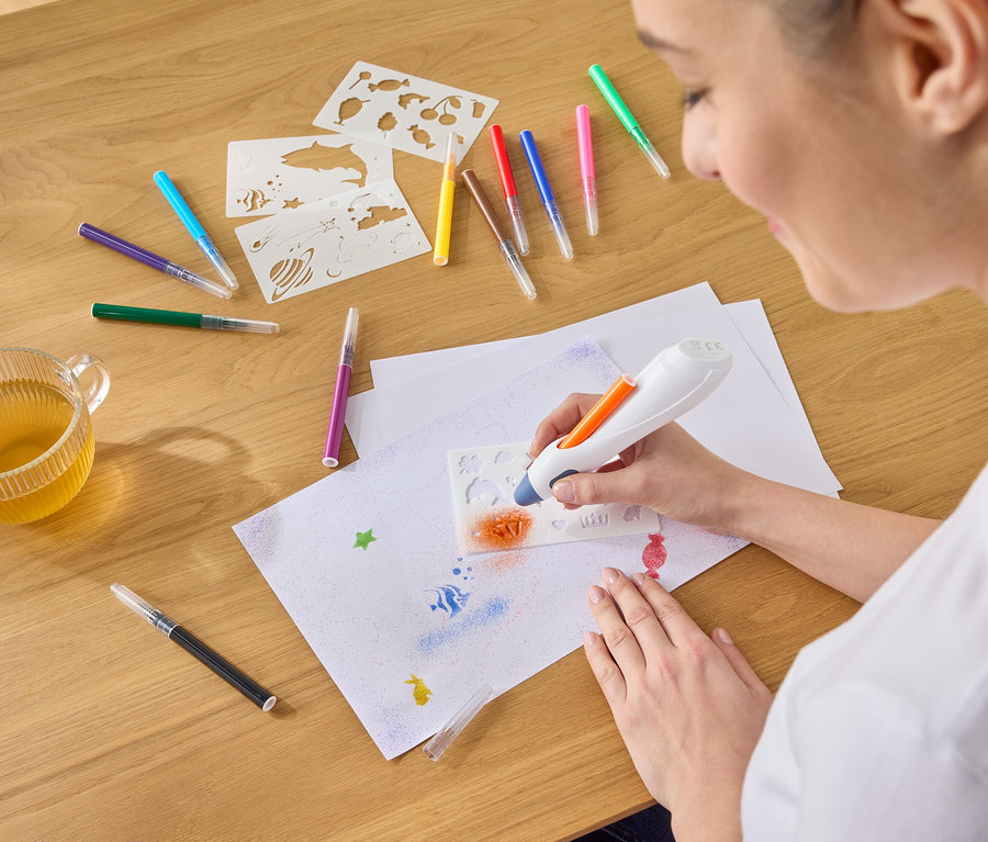 Une femme pulvérise de la peinture orange à l'aide d'un aérographe à travers un pochoir sur une feuille de papier, entourée de stylos, de pochoirs et d'une tasse de thé.