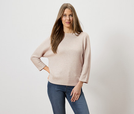 Une femme aux longs cheveux bruns, portant un pull rose clair et un jean, pose avec une main sur la hanche.