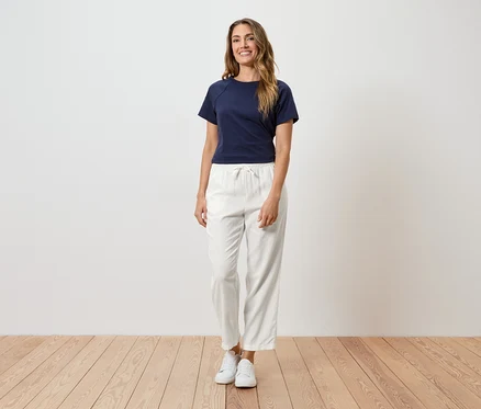 Une femme aux longs cheveux bruns ondulés portant un t-shirt bleu marine, un pantalon blanc à cordon et des baskets blanches.