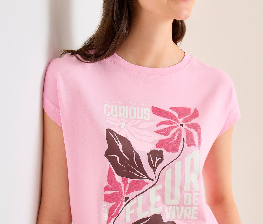 Femme avec une casquette beige et un T-shirt à motif CECIL rose avec un motif floral et l'inscription "FLEUR DE VIVRE" appuyée contre un mur.