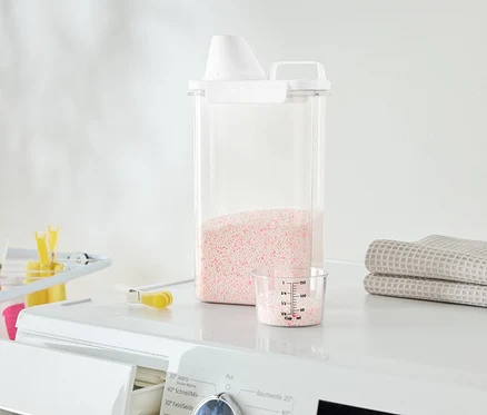 Sur un lave-linge se trouvent un récipient transparent avec de la lessive rose, un gobelet doseur et des serviettes pliées.