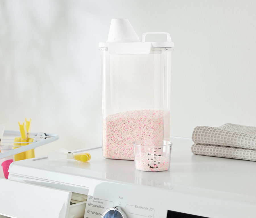 Sur un lave-linge se trouvent un récipient transparent avec de la lessive rose, un gobelet doseur et des serviettes pliées.