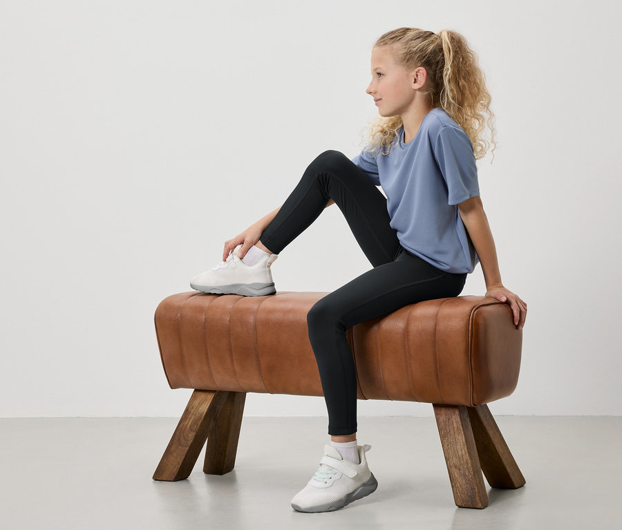 Une jeune fille aux cheveux blonds attachés en queue de cheval est assise sur un cheval d'arçons. Elle porte un t-shirt bleu, un legging noir et des baskets blanches.