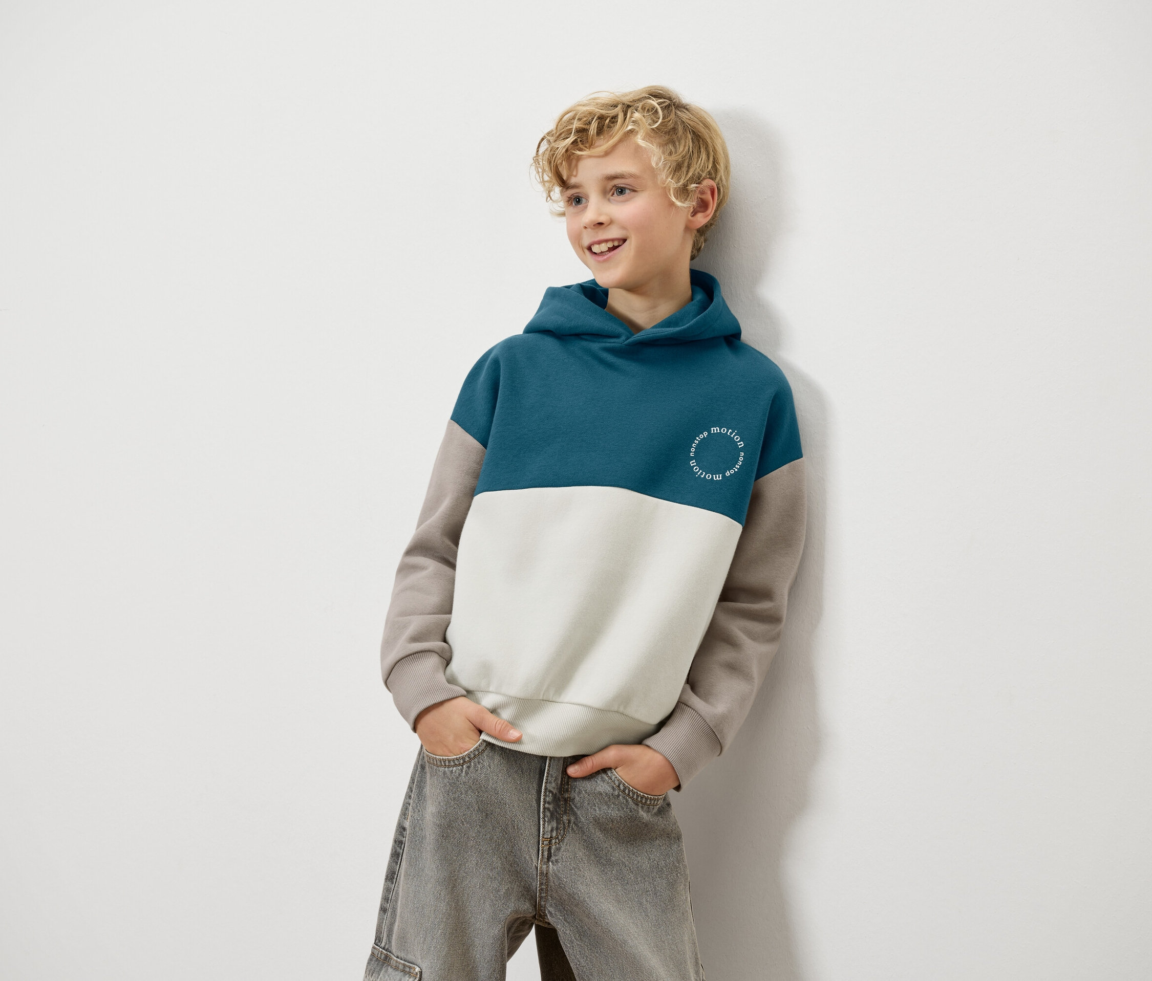Un garçon blond est appuyé contre un mur et porte un sweat à capuche bleu, blanc et marron et un jean gris.