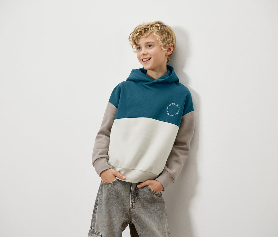 Un garçon blond est appuyé contre un mur et porte un sweat à capuche bleu, blanc et marron et un jean gris.