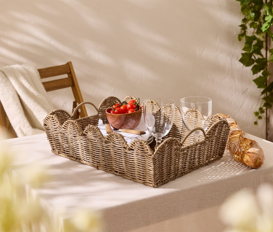 Sur une table avec une nappe se trouve un plateau tressé avec des tomates, des verres et une baguette.