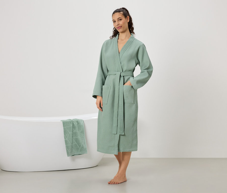 Une femme se tient pieds nus dans un peignoir vert menthe avec une ceinture et des poches, une main dans la poche. Une baignoire avec une serviette vert menthe est visible en arrière-plan.