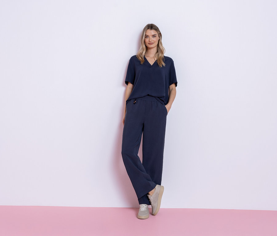 Une femme se tient devant un fond rose et blanc, portant une tenue bleu marine composée d'un t-shirt et d'un pantalon, ainsi que des baskets grises.